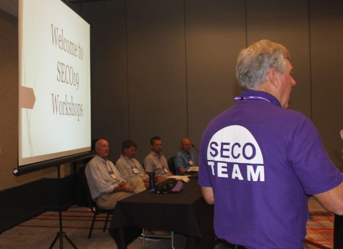 A presenter at SECO19 standing in front of a panel of speakers